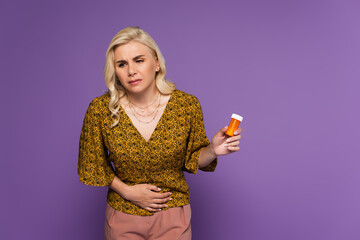 blonde woman with stomach ache holding bottle with pills isolated on purple.
