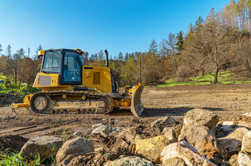 Obraz premium Side view working bulldozer clears the area before construction. Blue sky, forest background. Big stones.