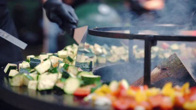 Traditional weekend cookout, man in gloves stirring cut vegetables with cooking tongs roasting on barbecue smoker grid. Green zucchini pieces, tomatoes, onion and bell peppers seasoned with aroma