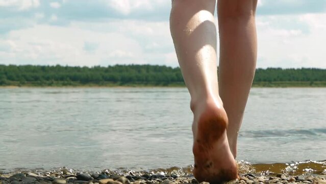 Red hair woman comes in water. Woman in red swimsuit comes in river on pebble bank. Closeup view on female feet on pebble river bank.