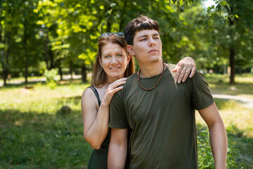 A woman and her son, who will soon be not a teenager but a young man, are chatting and having fun in the park. The boy is fooling around and the mother is proud of her son, smiles and hugs him