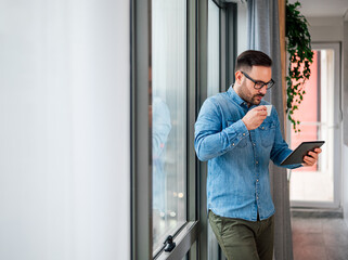 Young businessman or entrepreneur using digital tablet while enjoying coffee by the window at business office