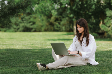 Beautiful Caucasian girl working with laptop computer sitting on green grass in summer park. Happy female outdoor working with technology, education and surfing the net, preparing for exams concept
