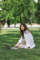 Beautiful Caucasian girl working with laptop computer sitting on green grass in summer park. Happy female outdoor working with technology, education and surfing the net, preparing for exams concept