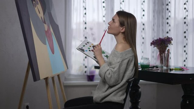 Side view gorgeous female painter making strokes with acrylic paint brush sitting at picture thinking. Talented inspired confident Caucasian woman enjoying hobby indoors. Creativity and fine art