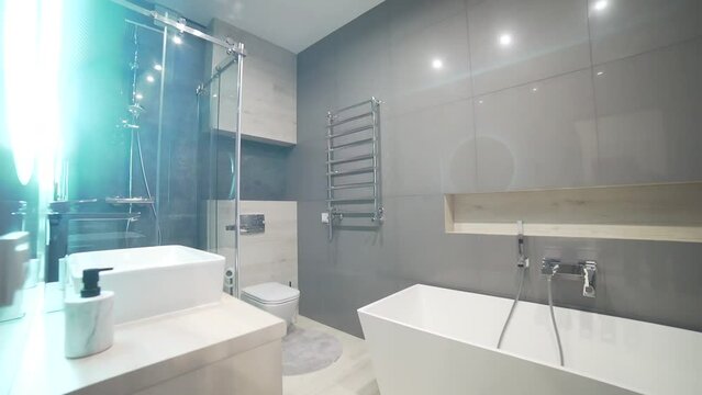 Video real interior a modern elegance bathroom in gray color with a white bathtub, a shower with a glass partition wall, a mirror with lighting and a toilet. in a house or apartment Steadicam shooting