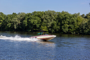 Fototapeta premium Modern speedboat sailing on forest background. Summer leisure and water activity. Rest On The River By Boat. Beautiful Motor Boat Rushes On The Water Surface Leaving Behind A Foam Trail