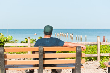 back view, medium distance of an adult male with a blue shirt and cap sitting on a wood bench at a tropical shoreline resting and relaxing