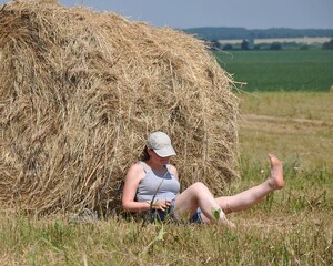 A woman sits in a field near a roll of hay. Raised bare foot. Hot summer day. Mowed grass. The person is resting after work on the field.