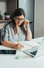 Smiling business woman sitting at kitchen desk and writing notes in a notebook while talking on a...