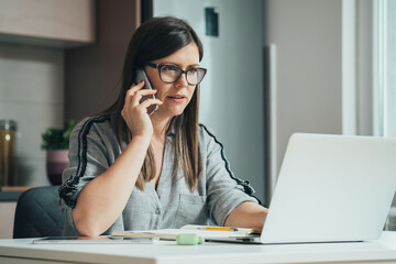 Serious Business Woman Working At Home On Her Laptop Computer While Talking On A Mobile Phone