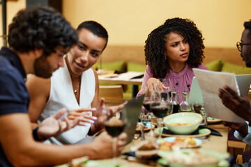 A group of multicultural managers is sitting in a restaurant at a dinner table, talking about a project and using technologies to improve it.