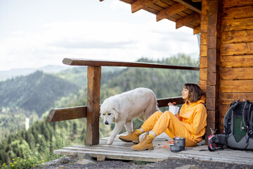 Young woman enjoys great mountain landscape and eats sublimated food for hiking, while sitting with her dog on a wooden terrace of nature house. Concept of solitude in nature and traveling with pets