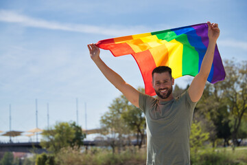 Young and handsome gay man, with a beard and green shirt, with blue eyes, perfect smile and waving a gay pride flag, smiling. Concept of gay pride, homosexual, lgtbi, pride day.