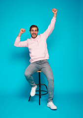 Full length of a cheerful young man sitting on chair and celebrating success isolated over light...