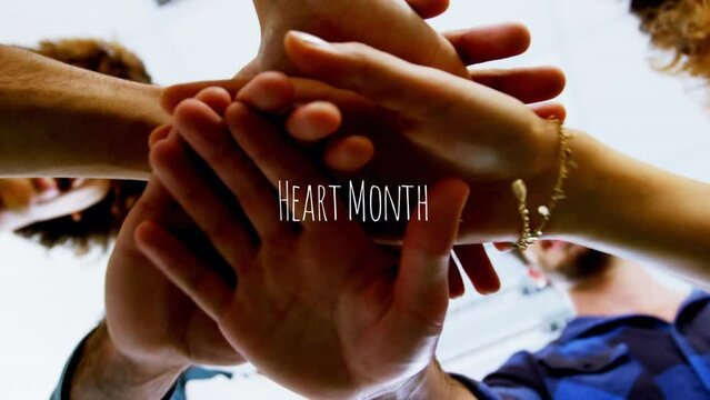 Heart month text over low angle view of diverse office colleagues staking hands together at office