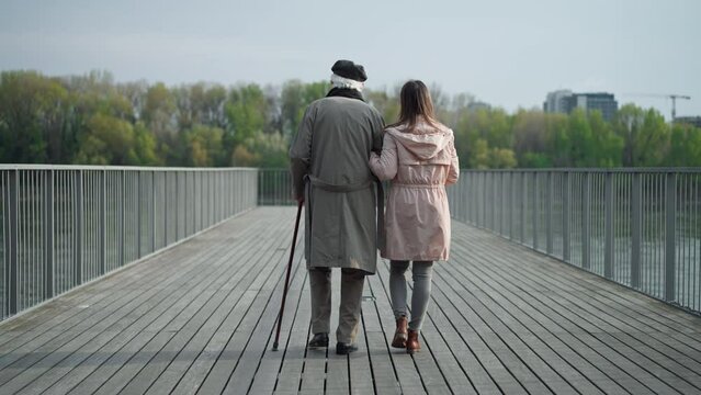 Rear view of senior man with daughter outdoors on a walk on pier by river.