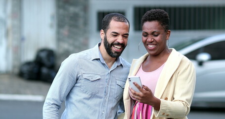Interracial couple laughing in street holding cellphone. Candid mixed couple relationship looking at smartphone. Real life laugh and smile