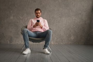 Cheerful Guy Using Mobile Phone With New Application Sitting In Armchair Over Gray Wall Background