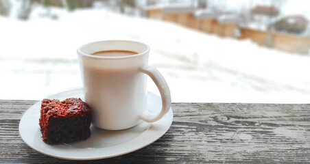 Winter frosty morning. A cup of coffee with milk and a piece of cake on a plate on a wooden table overlooking a snowy street