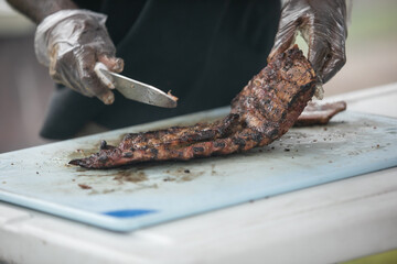 A man cutting up barbeque ribs from the smoker grill on cutting board with gloves on and a large knife.