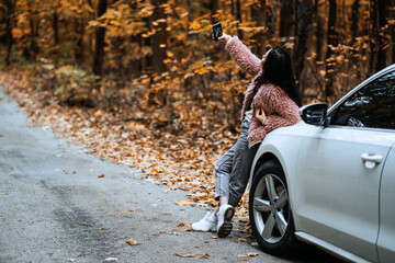 Confident brunette woman standing near car and doing selfie on cell phone. Fall autumn road weekend