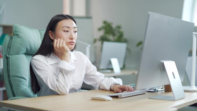 Bored sleepy Asian office worker sitting at work workplace at computer desk leaning on hand. Tired woman. Exhausted young Businesswoman employee. Portrait Depressed, unhappy, workless female manager