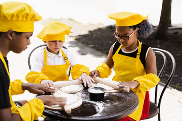 Multiethnic cooks children in yellow chefs hat and apron cooking dough for bakery. Black african and caucasian child cooking and having fun together.