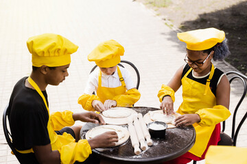 Multinational company of children cooks in yellow uniforms cooking dough for bakery. African teenager and black girl have fun with caucasian child boy and cook food.