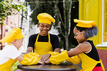 Teambuilding of multinational children cooks in chefs hat and yellow apron uniform put hands on each other, having fun and laughing. Multiethnic kids commutication activity.