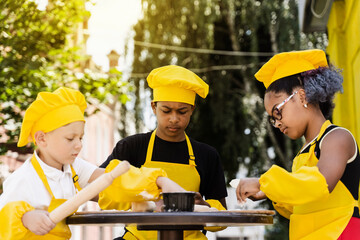 Multiracial cooks children in yellow chefs hat and apron cooking dough for bakery. Black african and caucasian child cooking and having fun together.