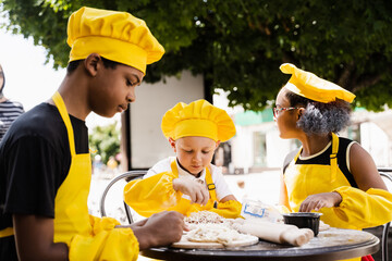 Childhood cook activity of multinational children of black african and caucasian kids in yellow chefs hat and apron uniform. Happy company of multiethnic children cooking dough.