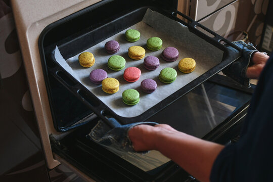 Woman hands taking out tray with multicolored macaroons pastry from the oven. Selective focus. 
