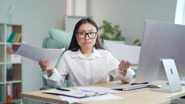 Difficult asian woman employee to deal with paperwork. documents. Stressed businesswoman confused by mistake, looking at camera through papers. bookkeeper, booker vaguely, dimly at work in office
