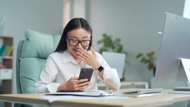 Asian business woman reading very funny news and messageson smartphone in office from smart phone, worker working in modern office. Employee watching a comic video in social networks at workplace