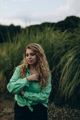 young beautiful woman with blond wavy hair posing near tall green grass. a plump girl in a green shirt and black trousers thought mysteriously about something.