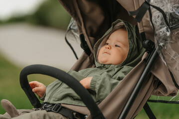 A female toddler is lying in the stroller on a cloudy day. A young girl in the raincoat is in her baby carriage in a park at noon.