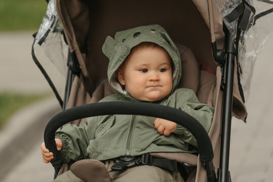 A female toddler is taking thought in the stroller on a cloudy day. A young girl in the raincoat is in her baby carriage in a park at noon.