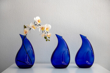 White artificial orchid in blue glass vase next to two empty blue glass vases on white table...