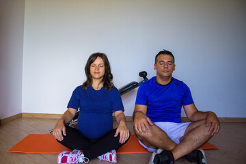 Image of a young pregnant woman with her husband in the home gym while doing meditation and yoga. Physical and mental preparation for childbirth