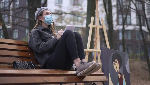 Focused young woman in coronavirus face mask sitting in autumn park drawing sketch. Talented inspired Caucasian abstract artist creating idea with contemporary picture leaned at bench