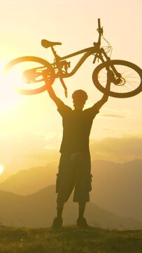 SLOW MOTION, SUN FLARE SILHOUETTE, COPY SPACE: Excited man lifts his bicycle overhead after a successful cross country biking journey in Slovenian mountains. Biker celebrates a great summer bike trip.