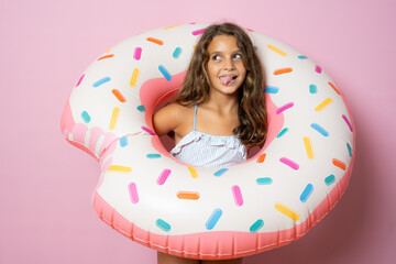 Obraz na płótnie Canvas Funny little brunette cute girl holding a big rubber ring on pink background. Active holidays with family on the beach pool. Emotional kid