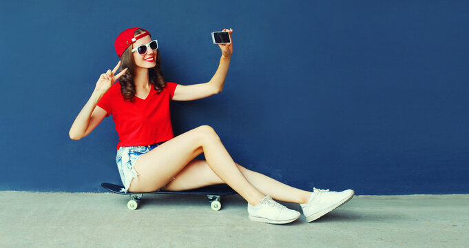 Happy smiling young woman taking selfie with smartphone sitting on skateboard wearing red baseball cap, shorts on city street on blue wall background
