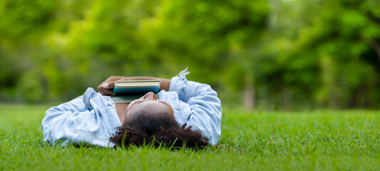 African American woman is lying down in the grass lawn inside the public park holding book in her hand during summer for reading and education concept with copy space