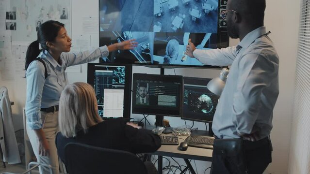 Blonde mature Caucasian woman sitting at desk, using computer in office at daytime, Hispanic detective and African American male analyst standing, looking at security cameras footage