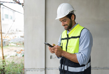 Young engineer in hardhat and safety vest typing text massage on mobile phone. Work environment of...