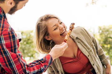 A young curvy woman laughs as she eats skewers of meat from her friend's hands during a trip to the...