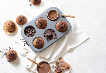 Top view of chocolate muffins flat lay in baking tray with slides of chocolate, chocolate chip, cocoa powder and chocolate sauce on white cutting board and white cloth