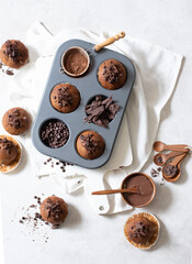 Top view of chocolate muffins flat lay in baking tray with slides of chocolate, chocolate chip, cocoa powder and chocolate sauce on white cutting board and white cloth